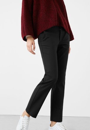 Person trägt schwarze Slim-Fit-Hose, weiße Sneakers und einen strukturierten dunkelroten Pullover, steht vor einem schlichten weißen Hintergrund.