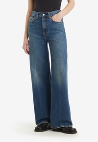 Personne portant un jean bleu taille haute à jambes larges, main dans la poche, associé à des chaussures noires et un haut noir sur fond blanc.