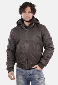 Giubbotto bomber in pelle marrone con cappuccio, zip frontale, polsini a coste e tasche. Indossato sopra una maglietta scura e jeans azzurri chiari.