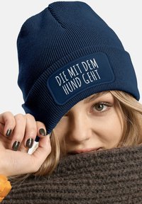 Navy blue knitted beanie with ribbed texture and a rectangular patch displaying white text. A hand partially holds the beanie.