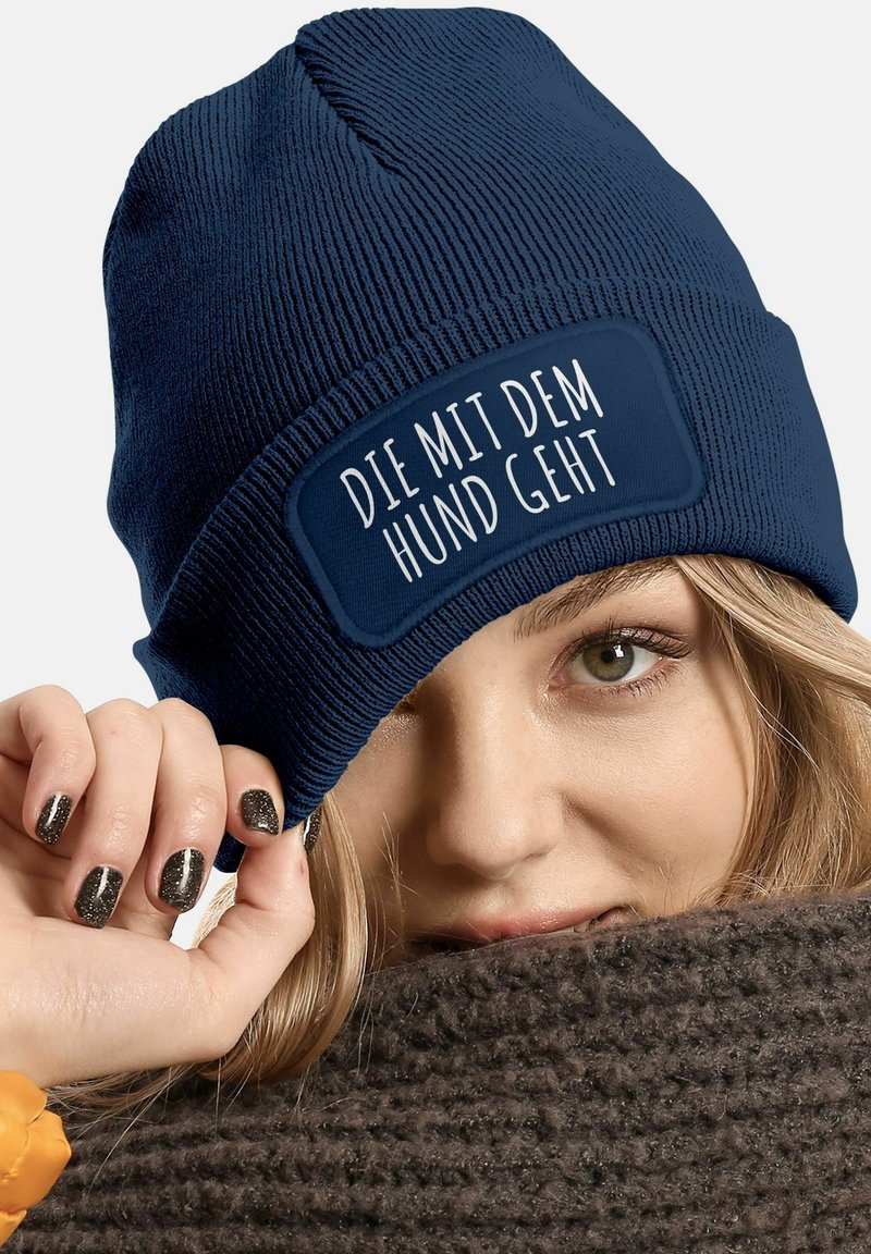 Navy blue knitted beanie with ribbed texture and a rectangular patch displaying white text. A hand partially holds the beanie.
