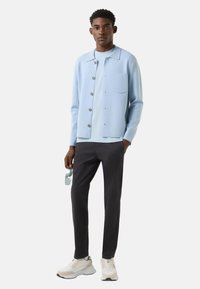 Světlomodrý cardigan na knoflíky s kapsou na hrudi, přes tričko ve stejném odstínu, doplněný tmavými kalhotami a bílými teniskami, držící sluneční brýle.