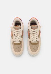 Béžové tenisky s oranžovými akcenty, svrchní část z texturované semiše a síťoviny, děrovaná špička a bílá gumová podrážka. Mají ploché tkaničky a značkový design.