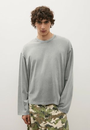 Jeune homme aux cheveux bouclés, portant une chemise grise ample à manches longues et un pantalon camouflage vert sur un fond clair uni.