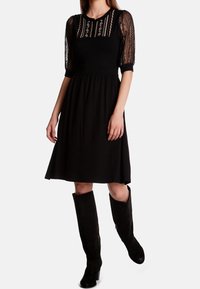 Robe noire avec des manches en dentelle, des broderies au niveau du col, et une taille cintrée s'évasant en jupe. Portée avec des bottes noires hautes.