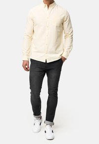 Mand iført en beige langærmet skjorte med knapper, sorte opbrettede skinny jeans og hvide sneakers, går med den ene hånd i lommen.