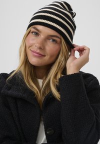 Berretto jacquard a strisce nere e crema con una texture a coste, indossato da una persona in un cappotto di pile nero, con una maglia liscia di colore neutro sotto.