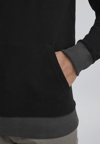Felpa nera con tasca frontale. Polsini e orlo a coste grigi. Texture del tessuto morbida. Dettagli di cucitura discreti. Vestibilità casual.