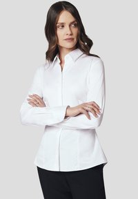 Biała koszula z długim rękawem, zapinana na guziki, o dopasowanym kroju, z klasycznym kołnierzykiem i gładką fakturą. Noszona z czarnymi spodniami.