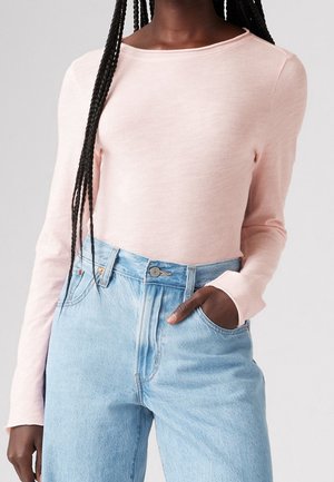 Persoon draagt een lichtroze shirt met lange mouwen, ingestopt in een high-waisted lichtblauwe spijkerbroek, met een hand in de zak.