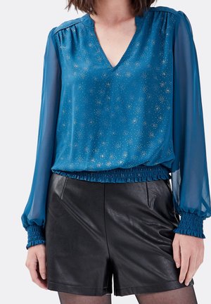 Femme portant un chemisier bleu transparent à manches longues avec des taches dorées et un short en cuir noir, debout devant un fond uni.