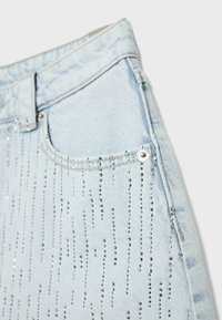 Poche de jean en denim bleu clair avec des rangées verticales de petits strass argentés et une rivet argentée près du bord de la poche.