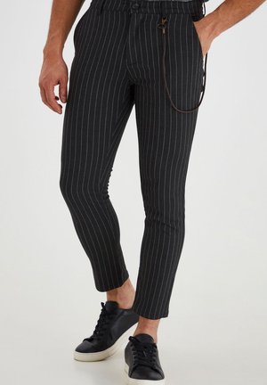 Mann trägt schwarze, schmal geschnittene Nadelstreifenhose mit Knöchellänge und schwarze Sneakers mit weißen Sohlen.