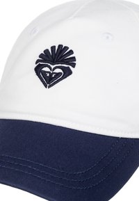 Gorra de béisbol blanca con visera azul marino, que presenta un diseño bordado en azul oscuro que se asemeja a un pájaro en la parte frontal. Detalles cosidos en la visera.
