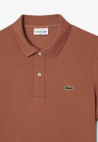 Brązowa polo Lacoste o slim fit z teksturowanej tkaniny, z trzema guzikami z przodu oraz wyszywanym zielonym logo krokodyla po lewej stronie klatki piersiowej.