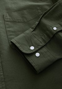 Tissu de chemise vert foncé avec un poignet replié et des boutons blancs. Présente une poche à soufflet et une surface texturée. Design structuré et décontracté.