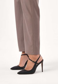 Czarne szpilki ze skóry ze spiczastym noskiem, z wąskim paskiem z tyłu i detalem klamry. Wysoki obcas typu szpilka i gładka faktura. W połączeniu z beżowymi spodniami.