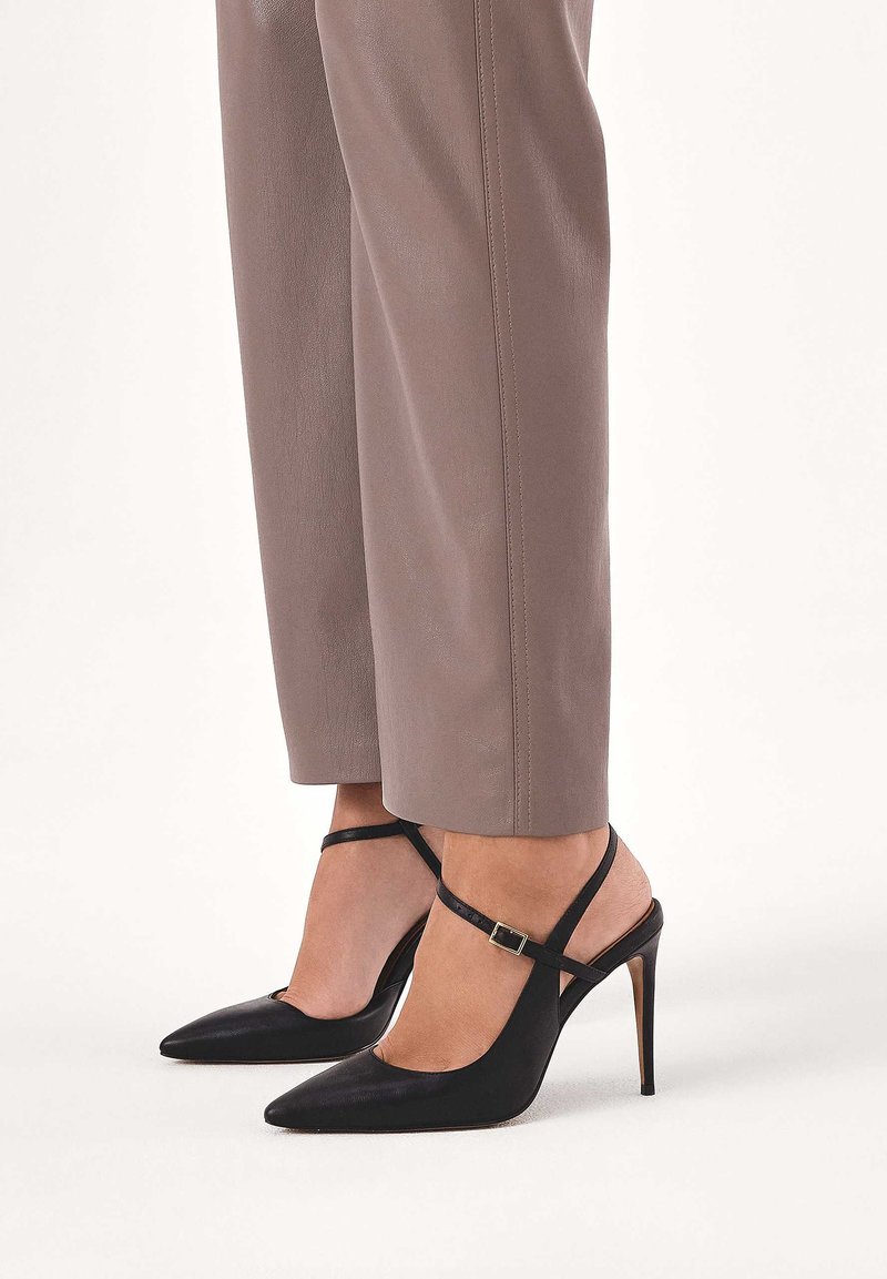 Czarne szpilki ze skóry ze spiczastym noskiem, z wąskim paskiem z tyłu i detalem klamry. Wysoki obcas typu szpilka i gładka faktura. W połączeniu z beżowymi spodniami.