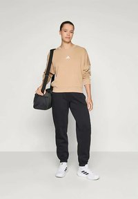 Beež kapuutsärk valge Adidas logo, mustad dressipüksid, valged sportlikud jalanõud mustade triipudega, kandes musta õlakotti.