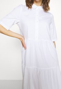 Abito in cotone bianco con maniche corte, chiusura a bottoni sul davanti e design a strati arricciati. Presenta un colletto e tasche laterali. Tessuto liscio.
