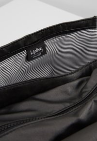 Intérieur d'un sac en tissu noir avec un motif à rayures, présentant une étiquette logo Kipling. Texture lisse et fermeture éclair visibles.