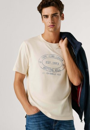 Beżowy bawełniany t-shirt z okrągłym niebieskim nadrukiem graficznym i tekstem, zestawiony z niebieskimi dżinsami i granatową kurtką.