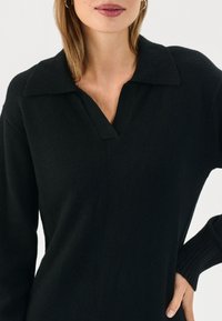 Pullover nero lavorato a maglia con scollo a V e colletto. Caratterizzato da maniche lunghe con polsini a costine, texture liscia e vestibilità rilassata.