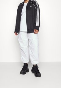 Mustakkainen vetoketjullinen treenijakku valkoisilla raidoilla, käytetty valkoisen t-paidan päällä ja yhdistetty löysiin valkoisiin housuihin sekä kömpelöihin mustiin lenkkareihin.