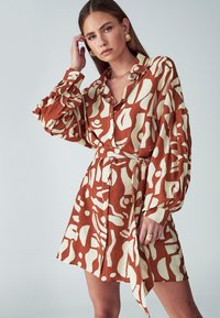 Sukienka koszulowa z lekkiego materiału w kolorze rdzawo-pomarańczowym z białymi abstrakcyjnymi wzorami. Posiada kołnierzyk, guziki oraz pasek.