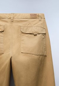 Back view of beige pants with two flap pockets and a small leather patch label on the waistband.
