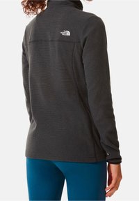 Fleecejacka i mörkgrått med hög krage, texturerat tyg och The North Face-logotyp på ryggen; ihop med blå leggings.