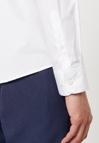 Detailní záběr na ruku osoby v bílém košili s knopovými manžetami, která je odložena podél těla, v kombinaci se strukturovanými tmavě modrými kalhotami.