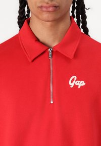 Crvena flis majica s patent zatvaračem, s bijelim izvezenim "Gap" logom na prsima. Glatka tekstura i klasičan dizajn.
