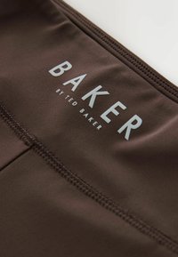Nahaufnahme eines dunkelbraunen Stoffes mit sichtbaren Nähten und weißem "BAKER BY TED BAKER"-Text, der darauf gedruckt ist.