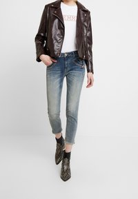 Burgundská kožená biker bunda, bílý grafický tričko, vybledlé modré skinny džíny s ozdobnými kapsami a černé kotníkové boty s hadím vzorem.