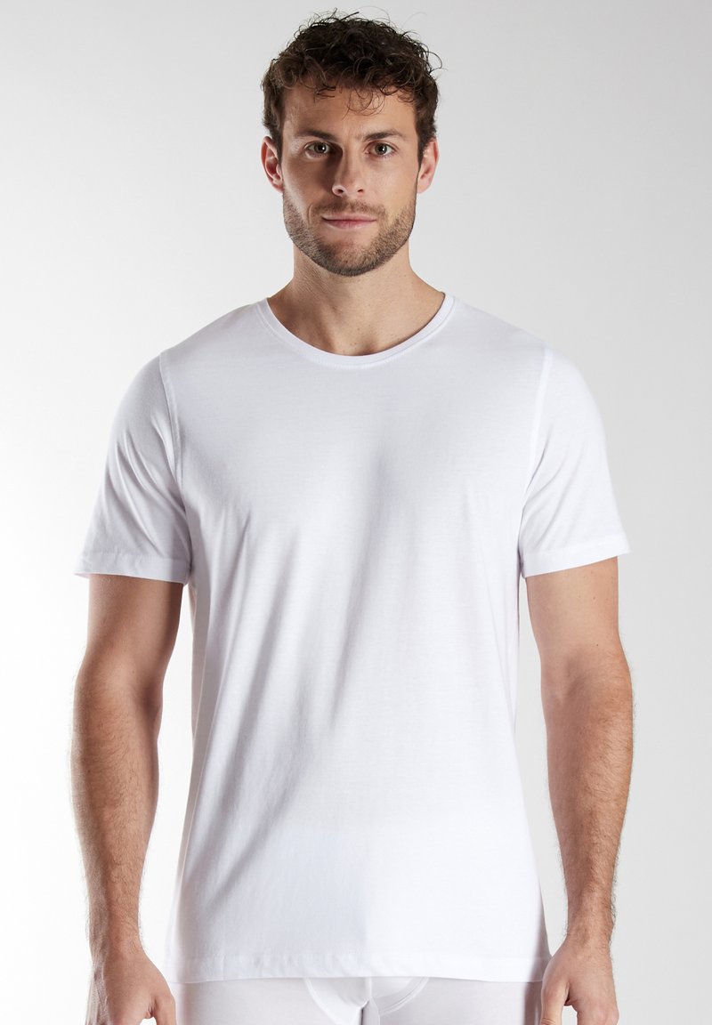 Man with short curly hair and beard wearing a plain white short-sleeve crew neck t-shirt standing against a light background.
