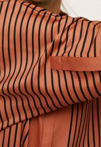 Nahaufnahme eines rostfarbenen langärmeligen Shirts mit schwarzen vertikalen Streifen und einer geknöpften Tab-Detaillierung am Oberarm.