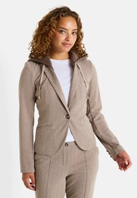 Blazer beige en chevrons avec capuche, doté de cordons de serrage et d'accents dorés, associé à un pantalon fuselé assorti. Chemise intérieure blanche visible.