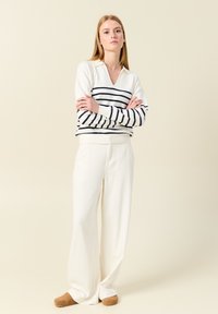 Weiß gestreifter Pullover mit V-Ausschnitt und langen Ärmeln, kombiniert mit hoch taillierten weißen weit geschnittenen Hosen und braunen Schuhen. Glatte Stoffoberfläche.