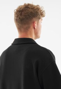 Fekete pamut ing gallérral, laza szabással és a háton látható varrási részletekkel, oldalról és hátulról készült felvételen.