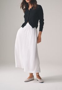 Sort cardigan med lange ærmer over en hvid, flydende maxinederdel. Beige netballerinaer med krydsede stropper. Bløde teksturer, kontrasterende farver.