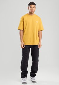 Mustard yellow cotton T-shirt with a crew neck and short sleeves, paired with black pants and white sneakers. Simple logo detail on the shirt.