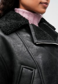 Κοντινό πλάνο ενός ατόμου που φοράει μια μαύρη δερμάτινη ζακέτα με γιακά από γούνα και ένα ροζ πλεκτό πουλόβερ με ψηλό λαιμό.