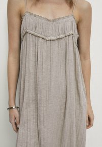 Robe sans manches, de couleur brun clair, fabriquée en tissu texturé. Elle possède un encolure froncée et des plis verticaux pour un design fluide.