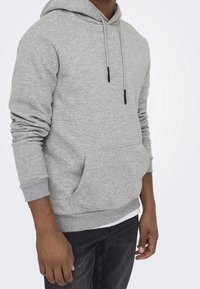Un sweat à capuche gris avec une poche avant et un cordon de serrage ajustable. Tissu doux et texturé avec des manches et un ourlet côtelés. Porté avec un jean foncé.