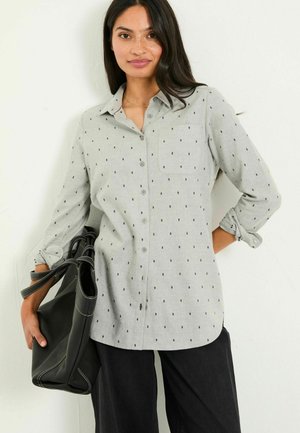 Graues Button-Up-Hemd mit schwarzem Punktmuster, langärmlig und an den Bündchen umgeschlagen, mit einer einzelnen Brusttasche, getragen mit einer schwarzen Tasche. Schwarze Hosen.