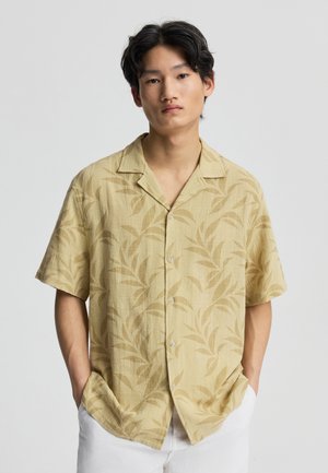 Giovane uomo con capelli neri che indossa una camicia beige a maniche corte con motivi di foglie e pantaloni bianchi, mani nelle tasche, sfondo neutro.