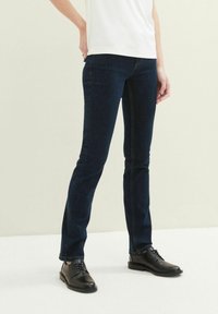 Jean en denim bleu foncé avec une coupe slim et un design classique à cinq poches, associé à des chaussures en cuir noires et un t-shirt blanc uni.