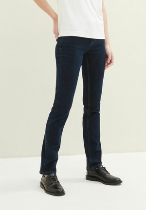 Jean en denim bleu foncé avec une coupe slim et un design classique à cinq poches, associé à des chaussures en cuir noires et un t-shirt blanc uni.