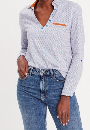 Femme portant une chemise à manches longues à motifs avec des boutons bleus et un détail orange, associée à un jean en denim bleu taille haute.